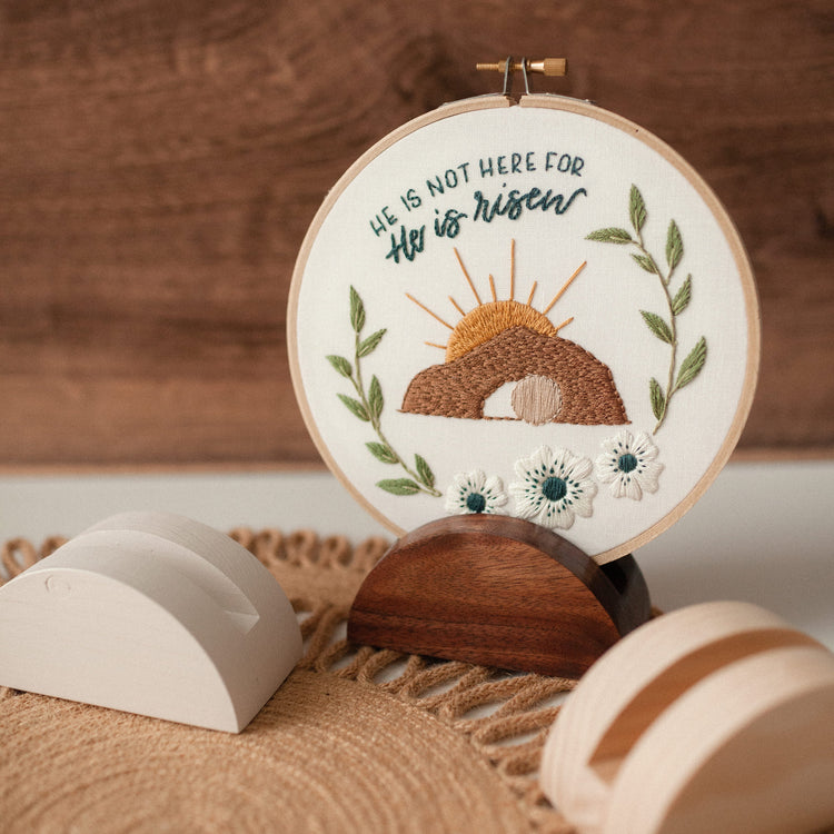 He Is Risen Easter embroidery hoop displayed on a walnut stand against warm wood