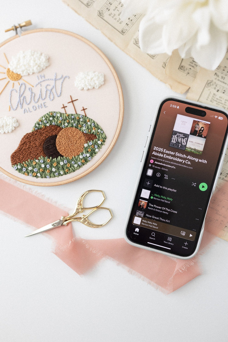 In Christ Alone hoop beside a phone showing the stitch-along playlist