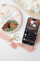 In Christ Alone hoop beside a phone showing the stitch-along playlist