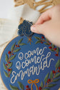 Close-up detail — hand placing the Glory ornament beside the finished hoop