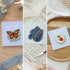 Three enamel pins displayed on light blue cards in a styled collage: a butterfly pin, a blue cursive word pin, and a strawberry flower pin. The cards are arranged on textured fabric, sheet music, and a wooden tray.
